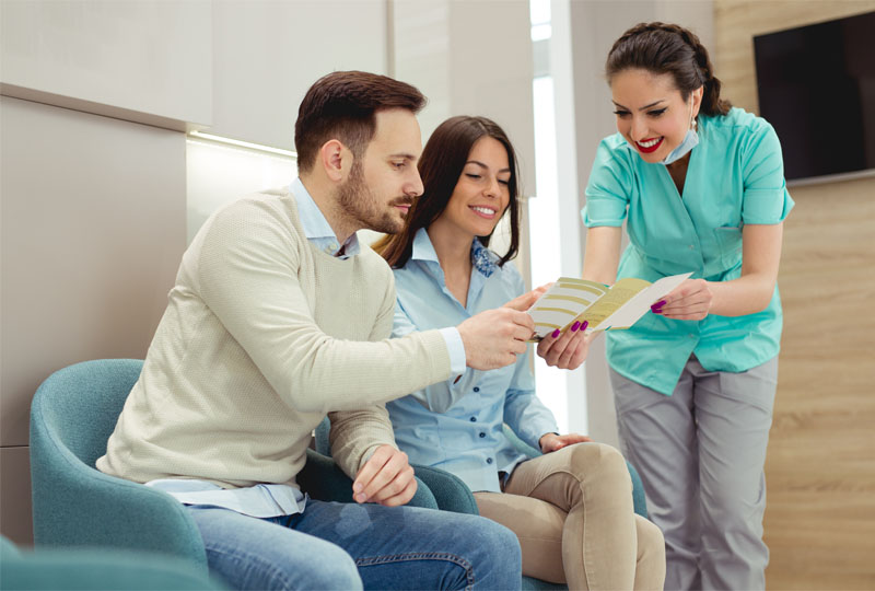 A photograph of three individuals, a man and two women, in a professional setting, seated on a couch with one woman holding an envelope while the others look at her.